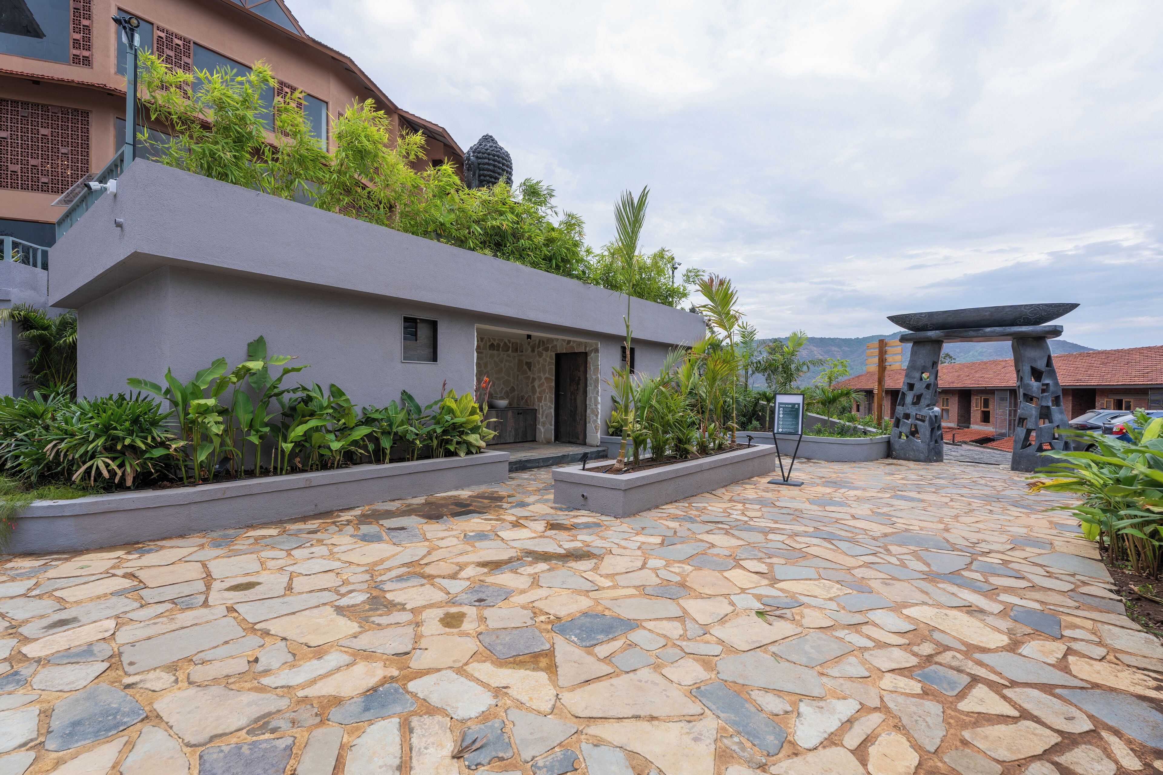 A stone-paved courtyard with a modern building, greenery, and a traditional stone structure.
