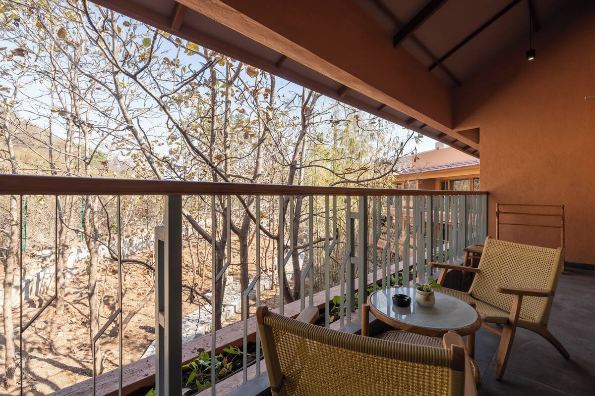 A balcony with wicker chairs and a small table, overlooking a dry landscape with trees and a building in the background.