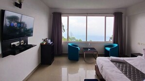 A hotel room with a bed, a TV, a desk, two chairs, and a large window with a view. - Veu Munnar Retreat (Devikolam)