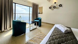 Quarto de hotel com cama, uma pequena mesa, uma cadeira, varanda com cortinas e luminária fixada na parede.