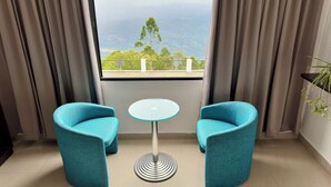 A room with two teal chairs, a small round table, and a view of a mountainous landscape through a large window. - Veu Munnar Retreat (Devikolam)