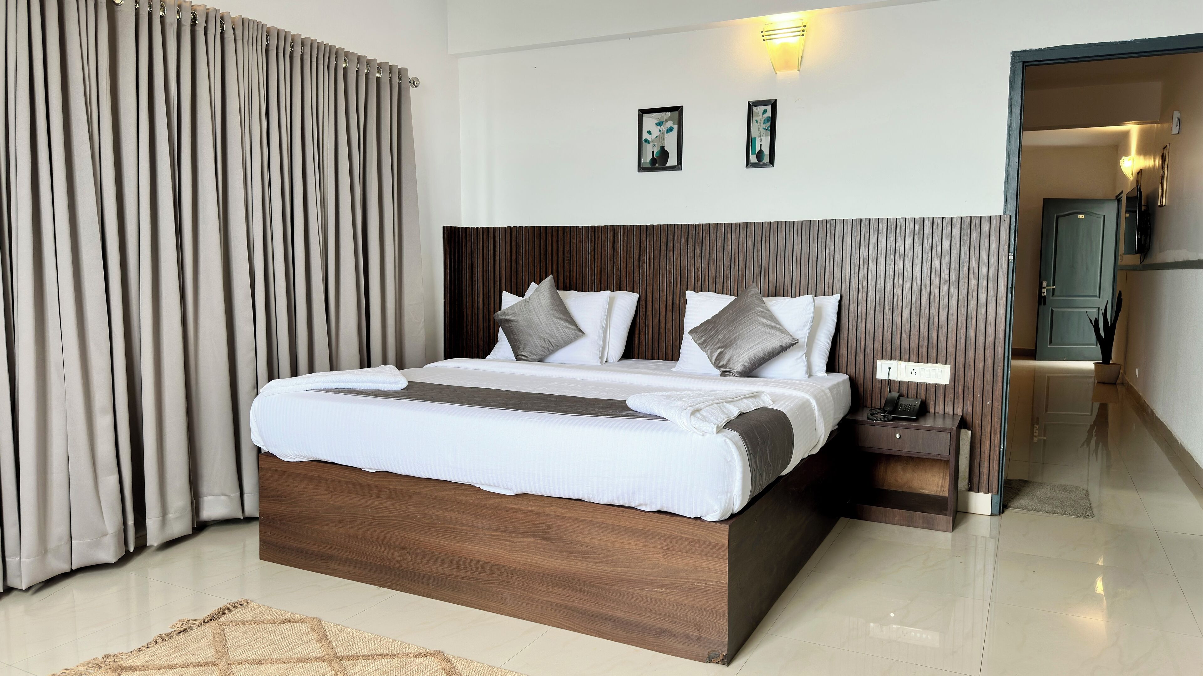 A modern hotel room with a wooden headboard, a large bed with white bedding, a bedside table with a phone, and a wall-mounted light fixture.