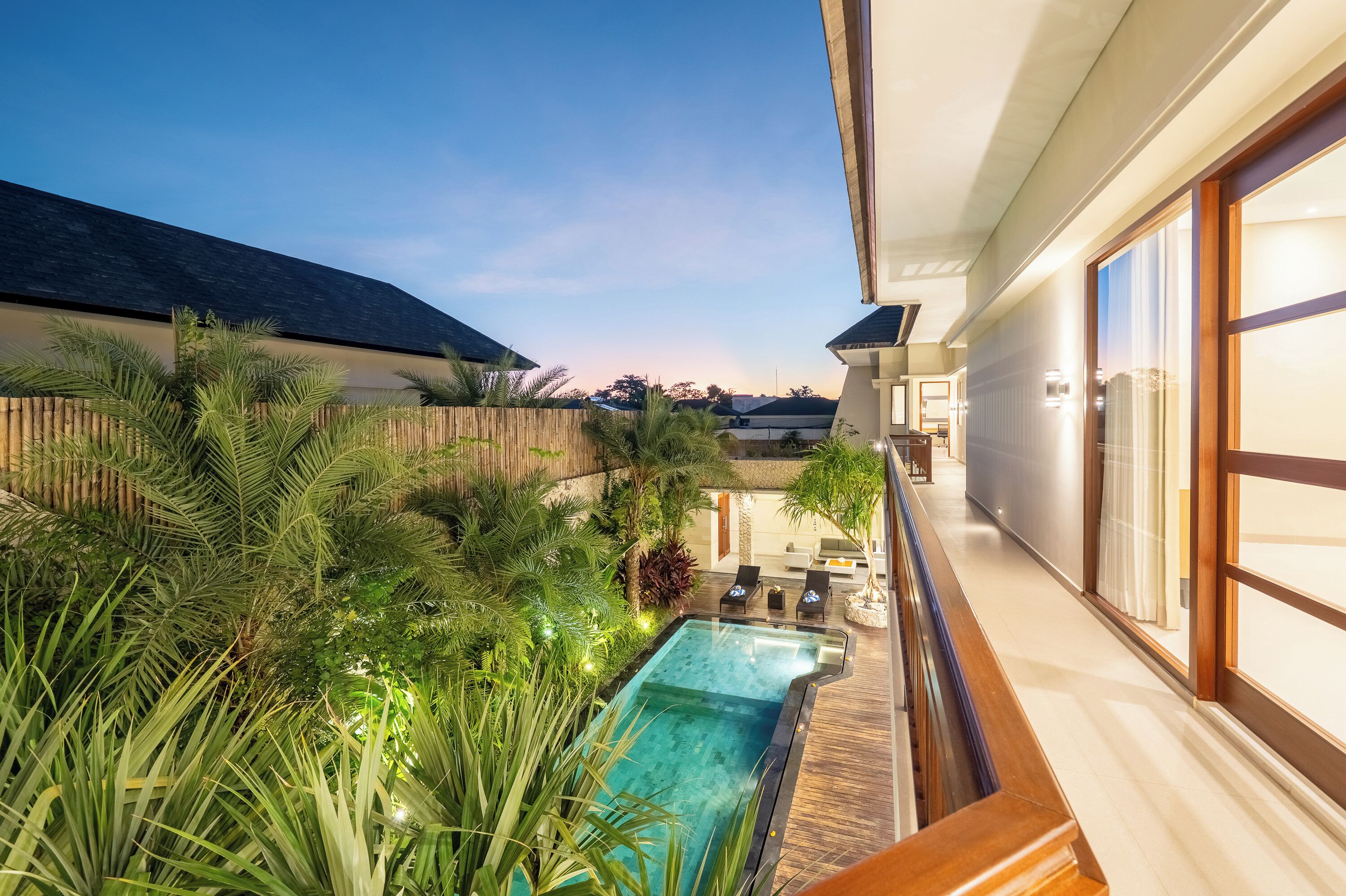 A modern outdoor pool area with a wooden deck, surrounded by lush greenery and palm trees.