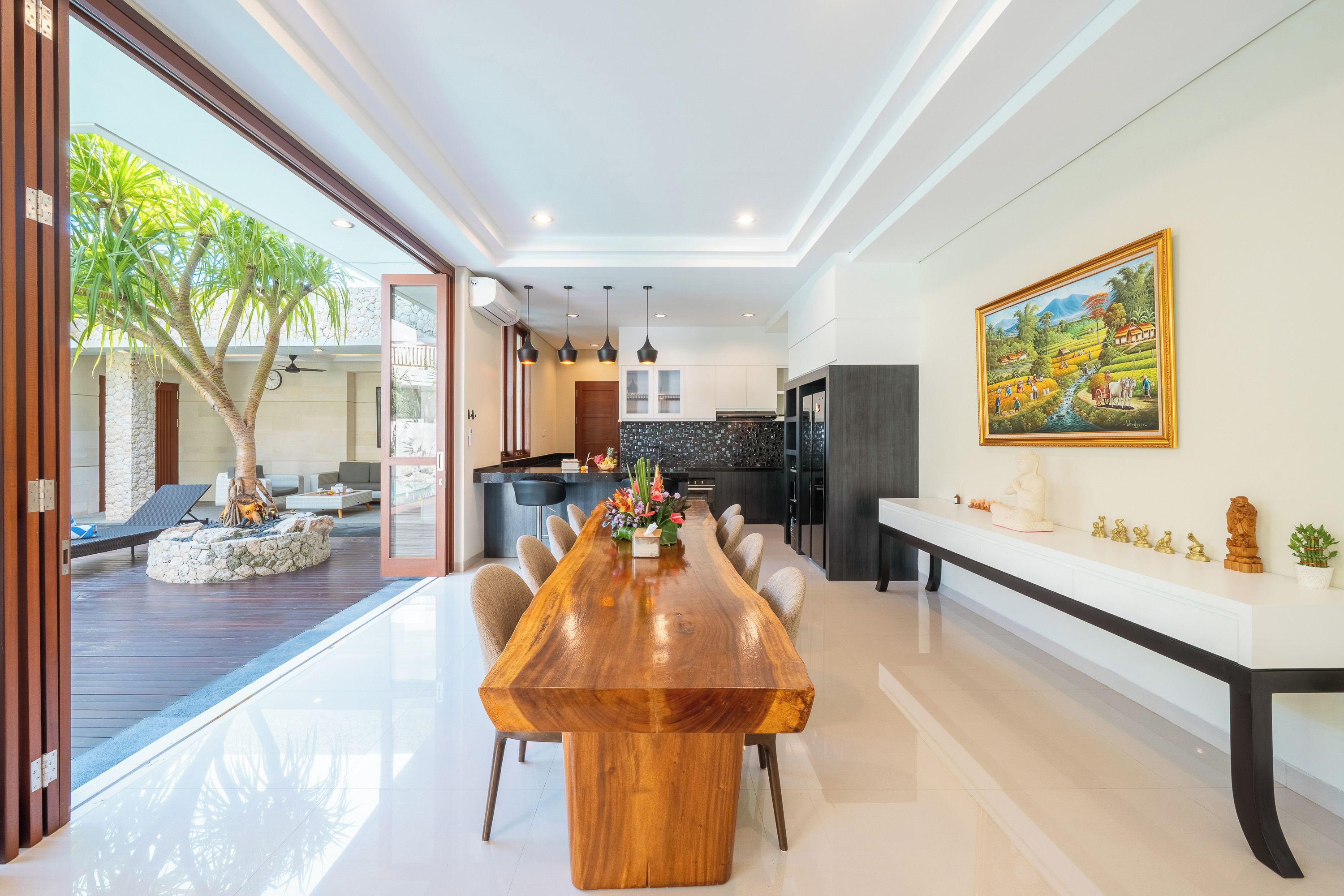 Un comedor amplio con una larga mesa de madera, rodeado de sillas, en una cocina moderna con una isla grande e iluminación colgante.