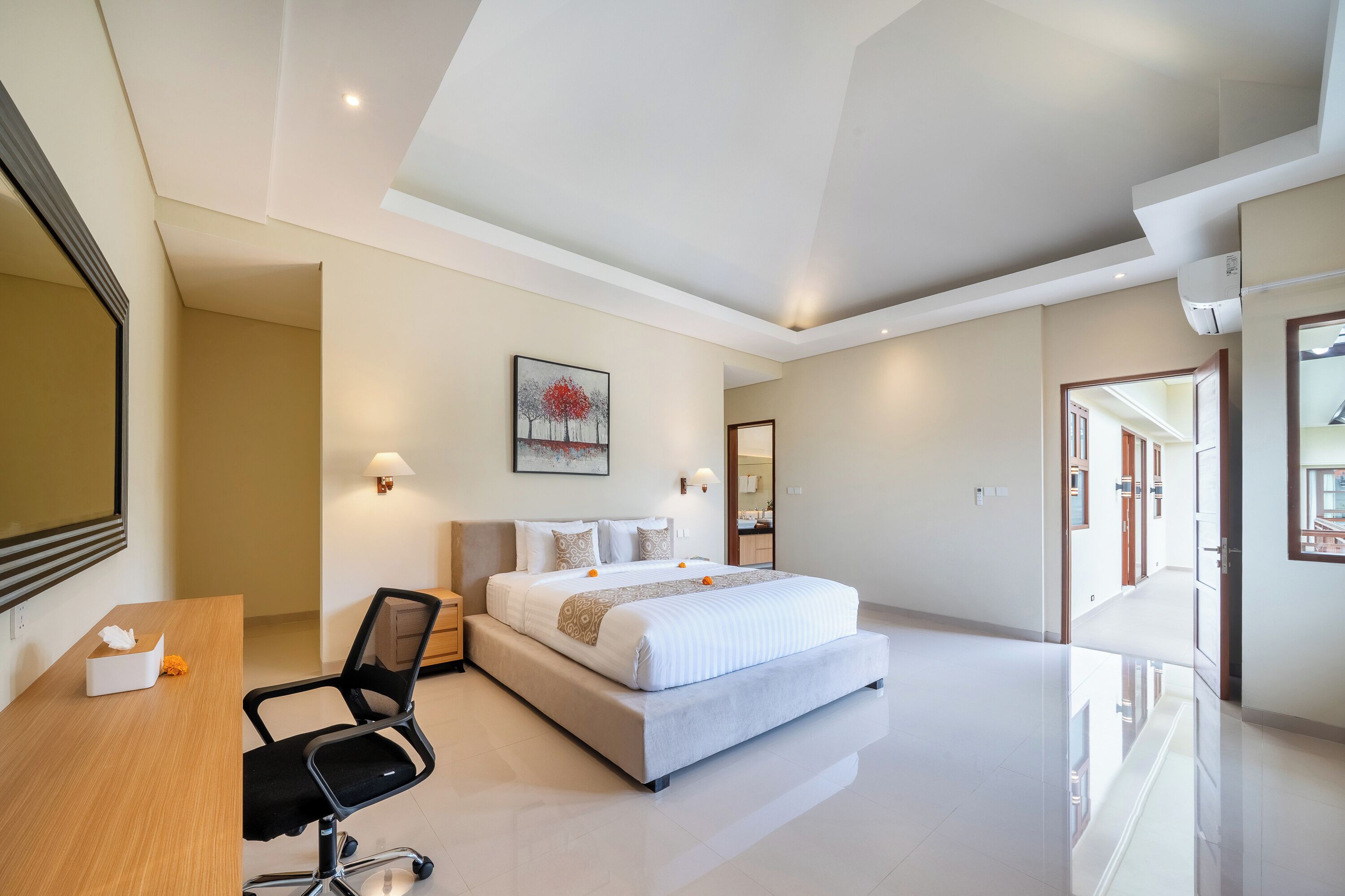 A modern hotel room with a large bed, a desk, and a chair. There is a painting on the wall and a mirror above the bed.