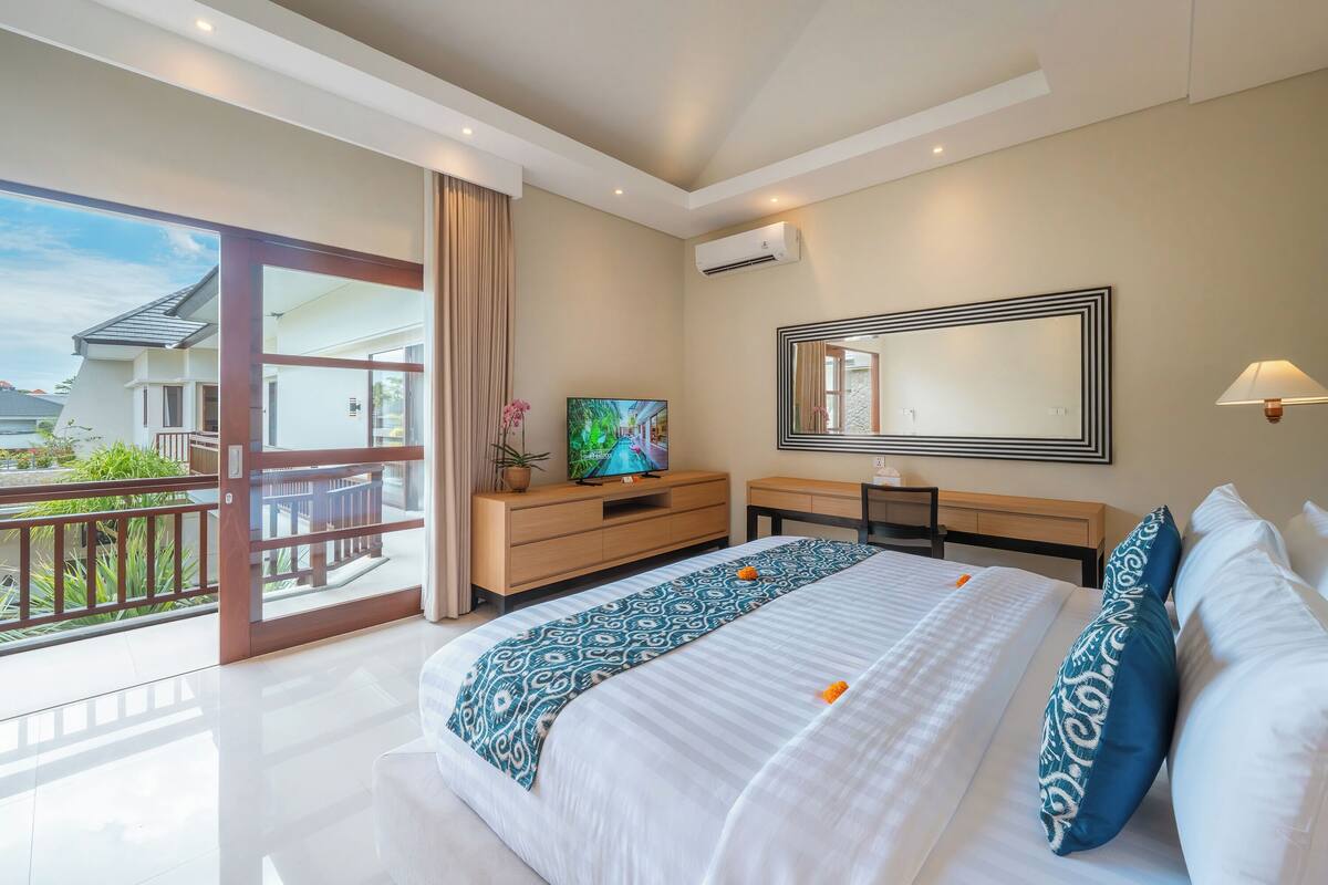 A hotel room with a large bed, a TV, a desk, and a balcony with a view of the surroundings.