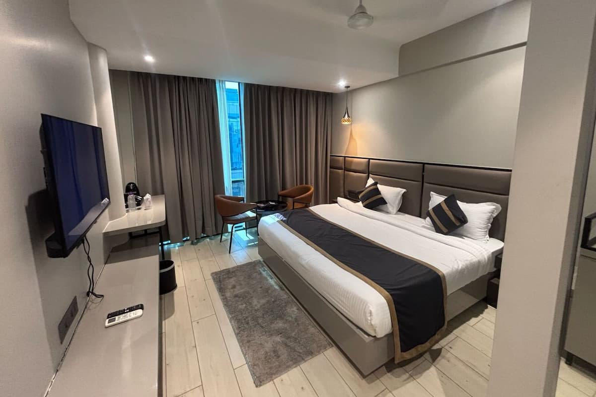 A modern hotel room with a large bed, a desk, a chair, a TV, and a view of the city.