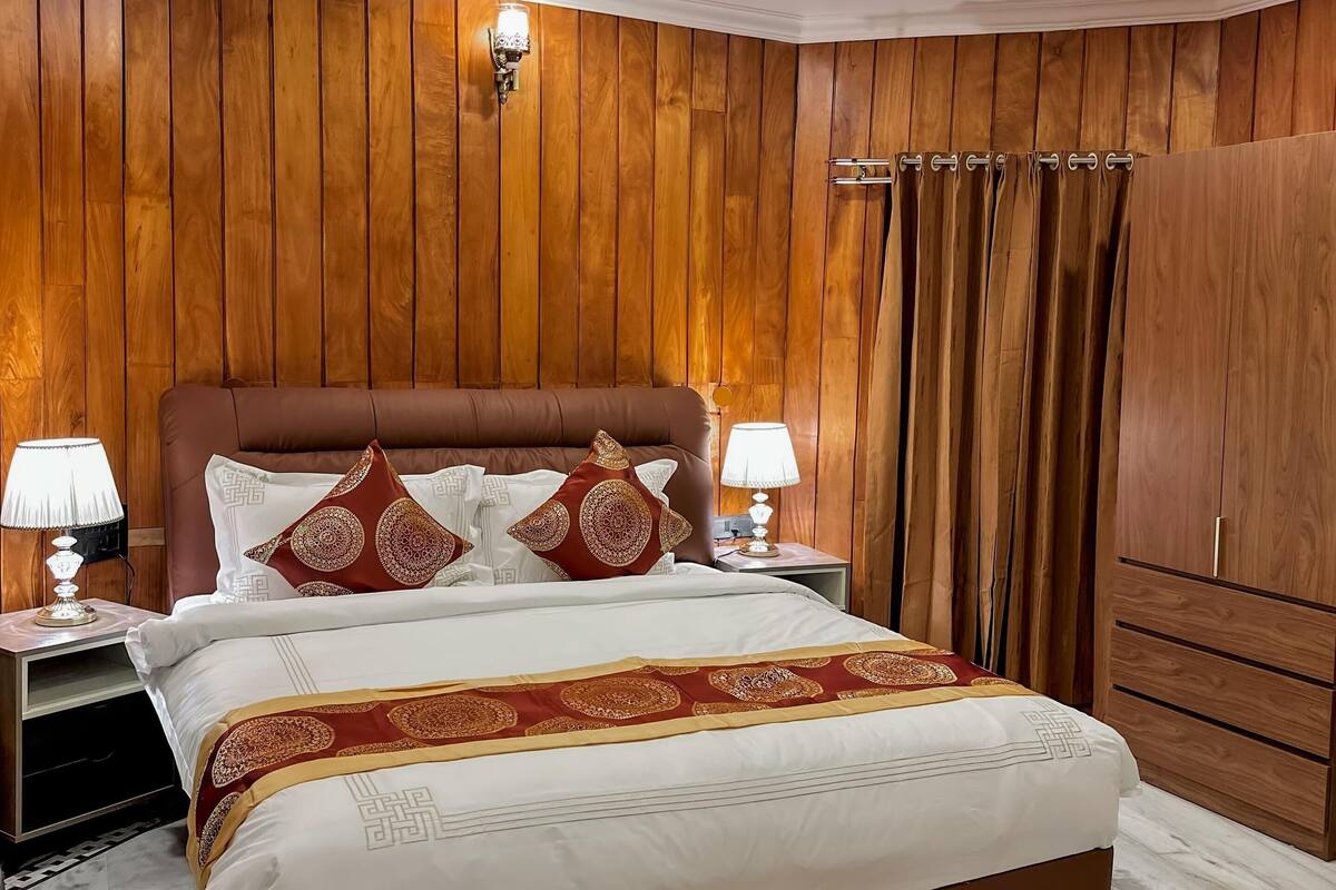Un dormitorio con pared de paneles de madera, una cama con colcha estampada, dos lámparas de noche y un armario.