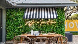 An outdoor dining area with a table set for four, surrounded by wicker chairs. A black and white striped awning provides shade, and a neon sign reads "Sizzle in San Antonio."