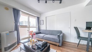 A modern living room with a grey sofa, a glass coffee table with decorative items, a standing heater, and a view of buildings through large windows. - SPACE 701 (Sapporo)
