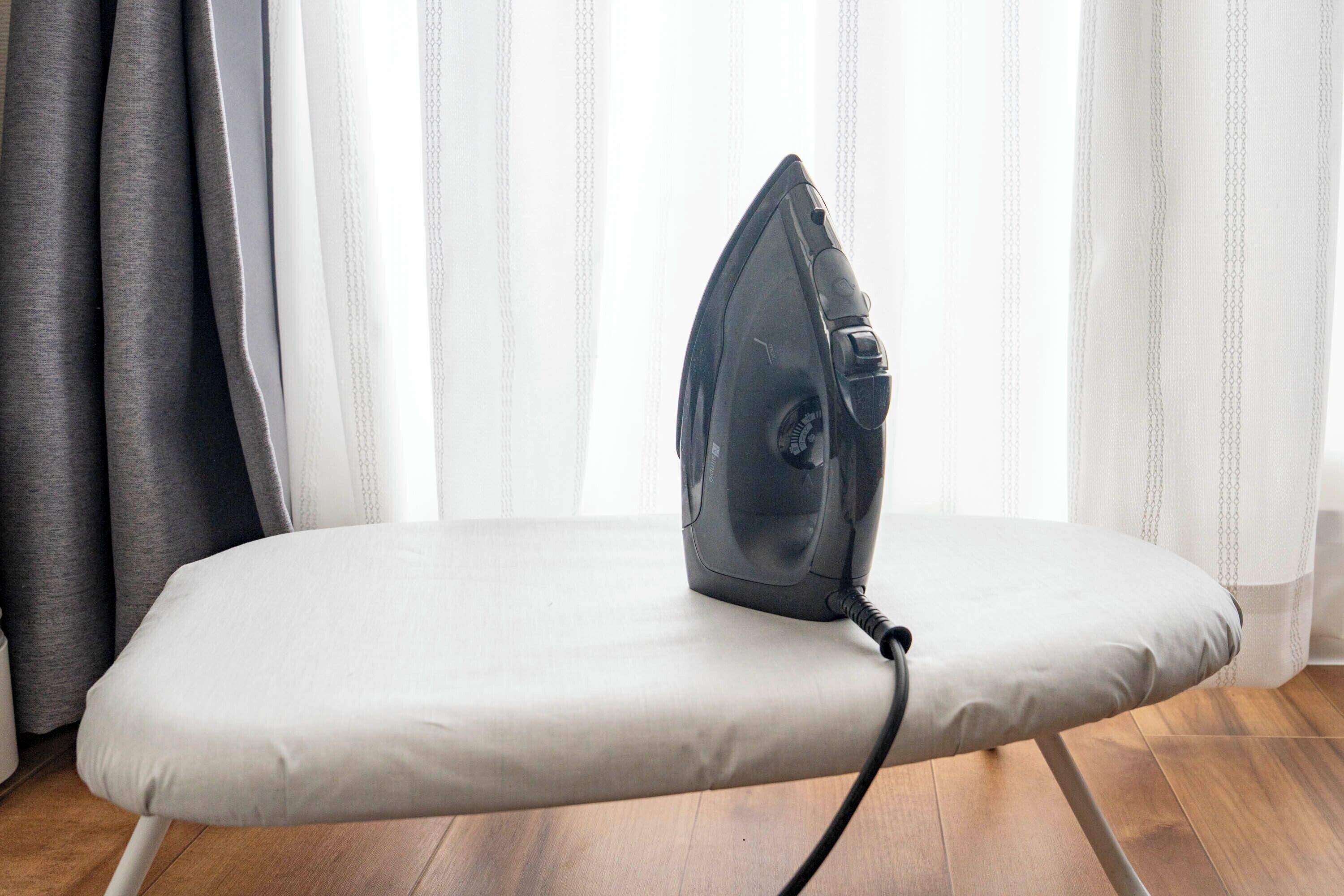 An ironing board with an iron placed on it, positioned in front of a window with curtains.