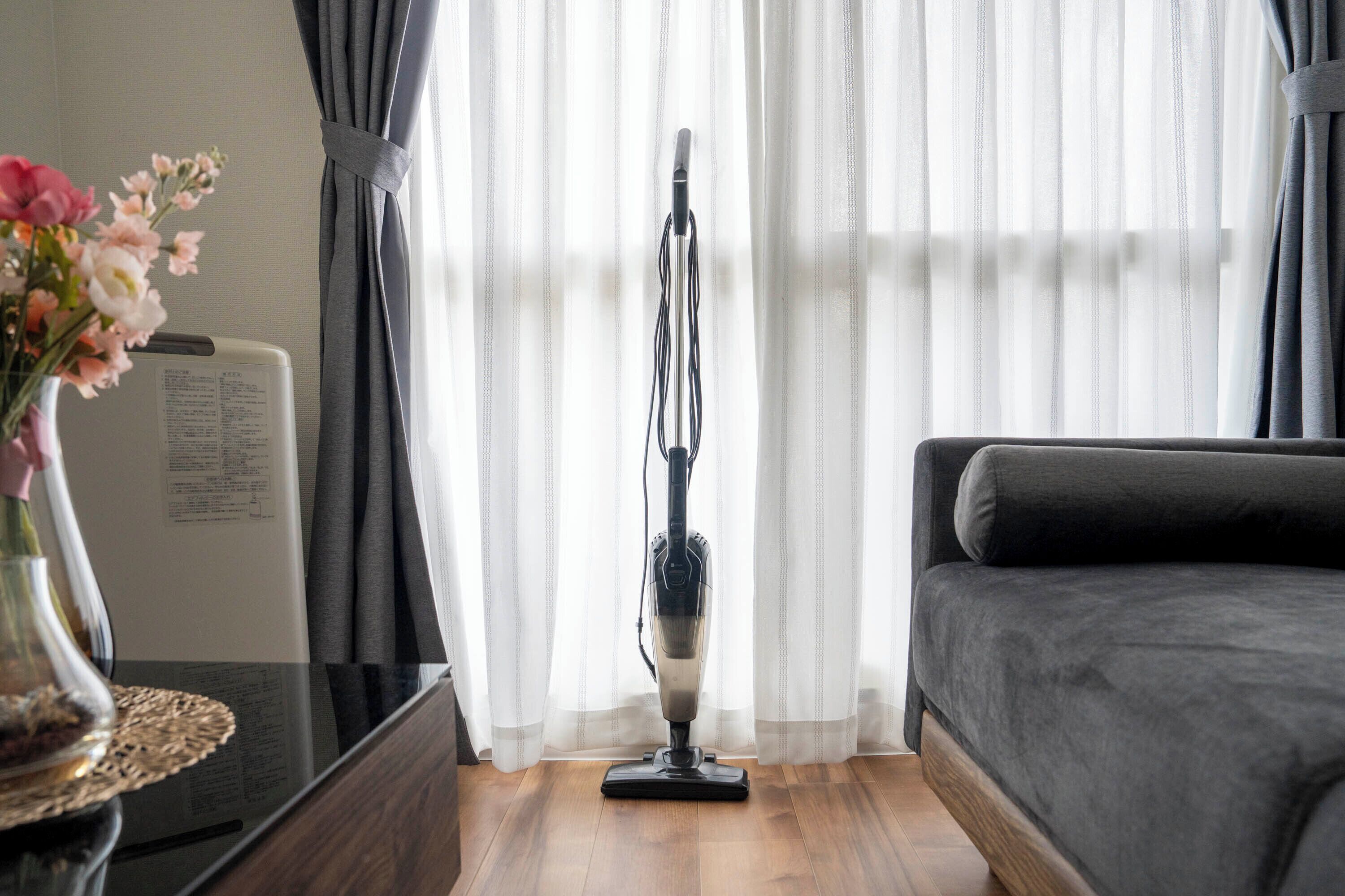 A room with a dark grey sofa, a standing vacuum cleaner, a wooden floor, and sheer curtains.