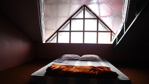 Une petite pièce à la lumière tamisée, avec un lit simple, une fenêtre de forme triangulaire et un plafond en pente.