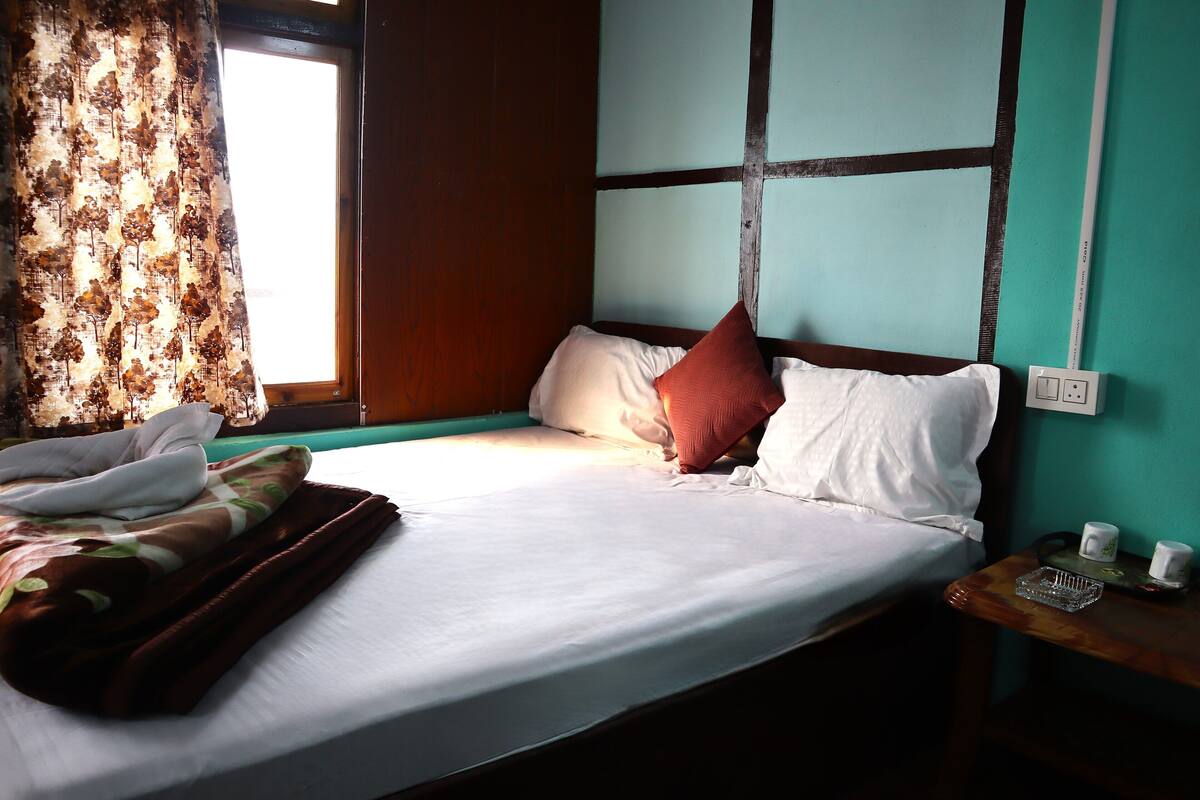 A neatly made bed with white linens and a brown blanket, a wooden nightstand with a tray and two cups, a window with curtains, and a wall with a grid pattern.