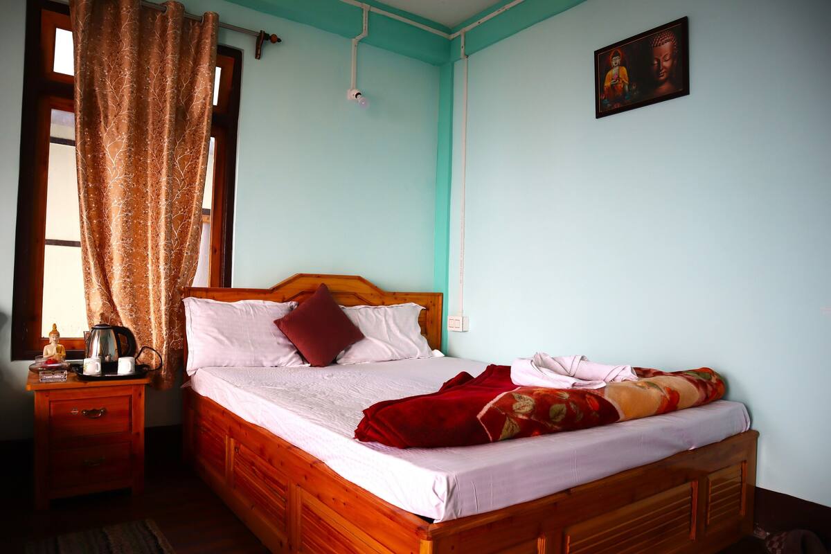 A neatly made bed with a wooden headboard, a bedside table with a kettle and cups, a window with curtains, and a wall-mounted picture.
