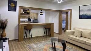 A modern living room with a kitchen area, a glass coffee table, and a sofa. - Blossom Service Apartment  (Lagos)
