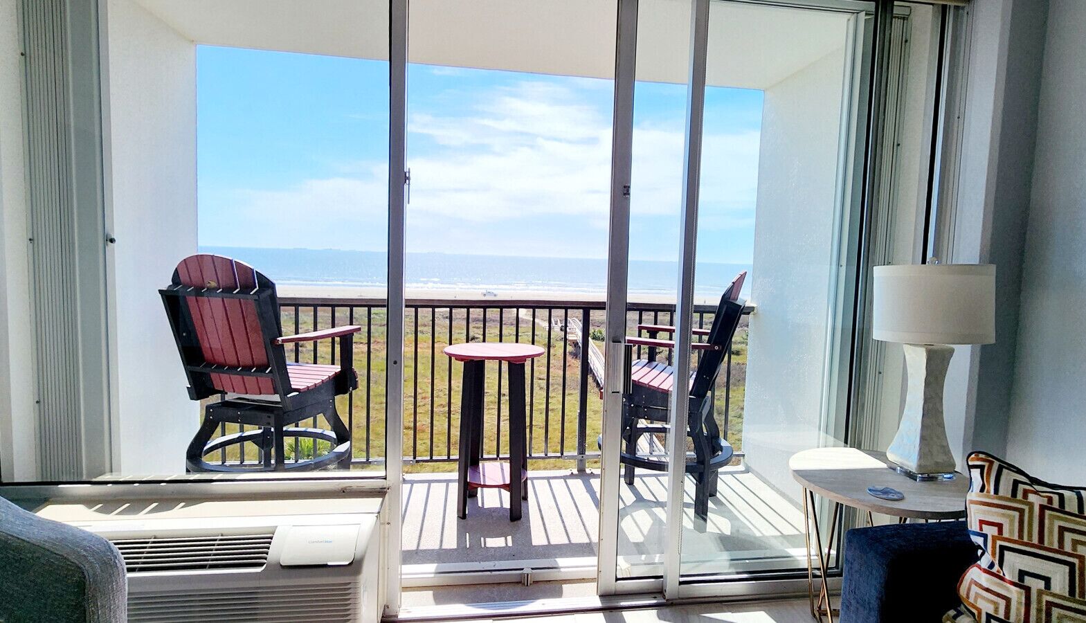 A balcony with a view of the ocean, a red chair, a small round table, and a lamp.