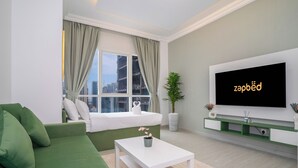 Een moderne woonkamer met een groene bank, een witte salontafel, een grote flatscreen televisie en een raam met gordijnen.
