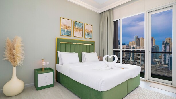 Een moderne hotelkamer met een groen bed, witte beddengoed, een nachtkastje met lamp, een vaas met pluimgras en een stadszicht door grote ramen.