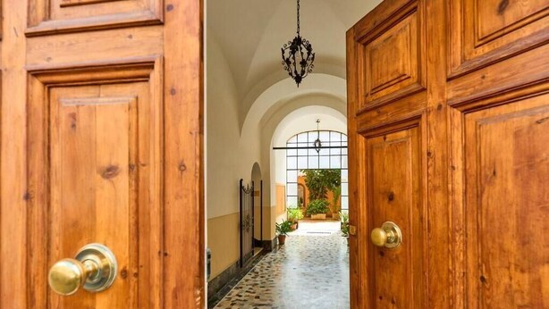 A wooden door with a brass doorknob, leading to a hallway with a tiled floor and a view of a garden through a window. - LABI HOME COLOSSEVM (Roma)