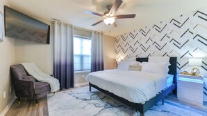 A bedroom with a bed, a chair, a ceiling fan, a TV, and a patterned wall. - Entire Home-Beautiful 4BR, 3.5BA Retreat in Ankeny, Iowa 2 King Beds, Fenced, AC (Ankeny)