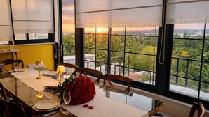 Un coin repas avec une table dressée pour deux personnes, un bouquet de roses rouges et une vue sur la verdure à travers de grandes fenêtres.