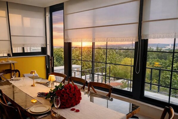 Un coin repas avec une table dressée pour deux personnes, un bouquet de roses rouges et une vue sur la verdure à travers de grandes fenêtres.