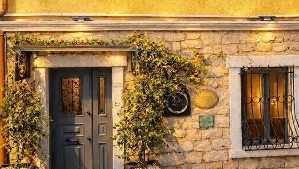 Un bâtiment à deux étages, avec une façade jaune, un rez-de-chaussée en pierre et une porte bleu foncé agréée d’un portail en métal.