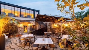 Une terrasse extérieure avec un sol en pierre, des tables et des chaises, un escalier et un bâtiment doté de grandes fenêtres en arrière-plan.