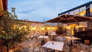 Un'area patio esterna con pavimento in pietra, tavoli e sedie, circondata da un muro in pietra e un edificio con ampie finestre.