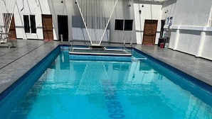 Une piscine rectangulaire à l’eau d’un bleu limpide, entourée d’une terrasse carrelée et bordée d’un bâtiment blanc doté de nombreuses portes et fenêtres.