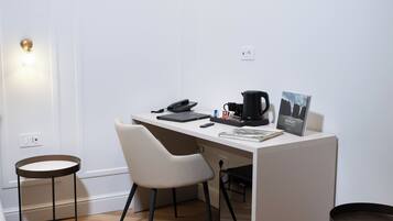 A small, modern workspace with a white desk, a laptop, a phone, a coffee maker, and a framed picture. There are two chairs and a small table.