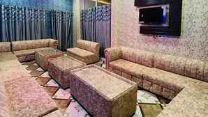 A modern living room with a sectional sofa, a flat-screen TV mounted on the wall, and a decorative chandelier. - Hotel Rajveer Inn (Varanasi)