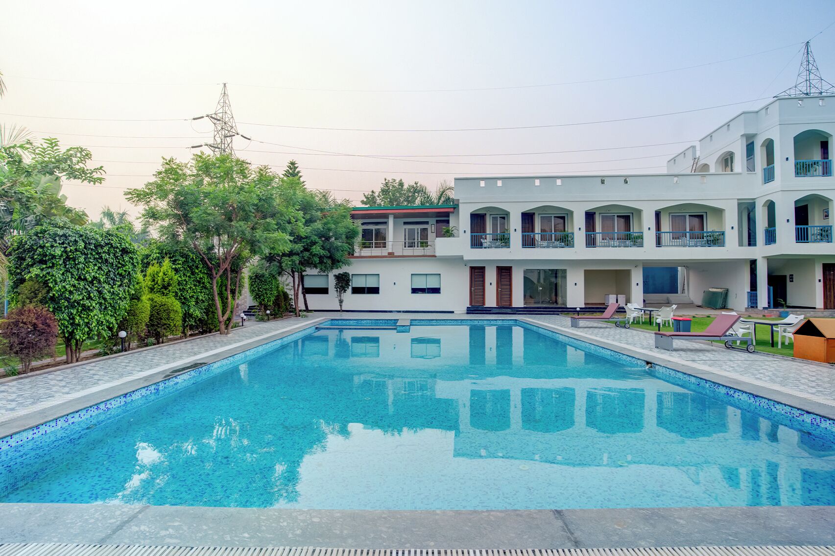 Ein Swimmingpool mit glasklarer, blauer Wasseroberfläche, umgeben von einer gepflasterten Liegefläche. Auf der Liegefläche stehen Liegestühle und ein kleiner Tisch. Im Hintergrund ist ein mehrstöckiges Gebäude mit Balkonen und Fenstern zu sehen.