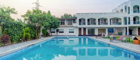 A swimming pool with a clear blue water surface, surrounded by a paved deck. There are lounge chairs and a small table on the deck. In the background, there is a multi-story building with balconies and windows.