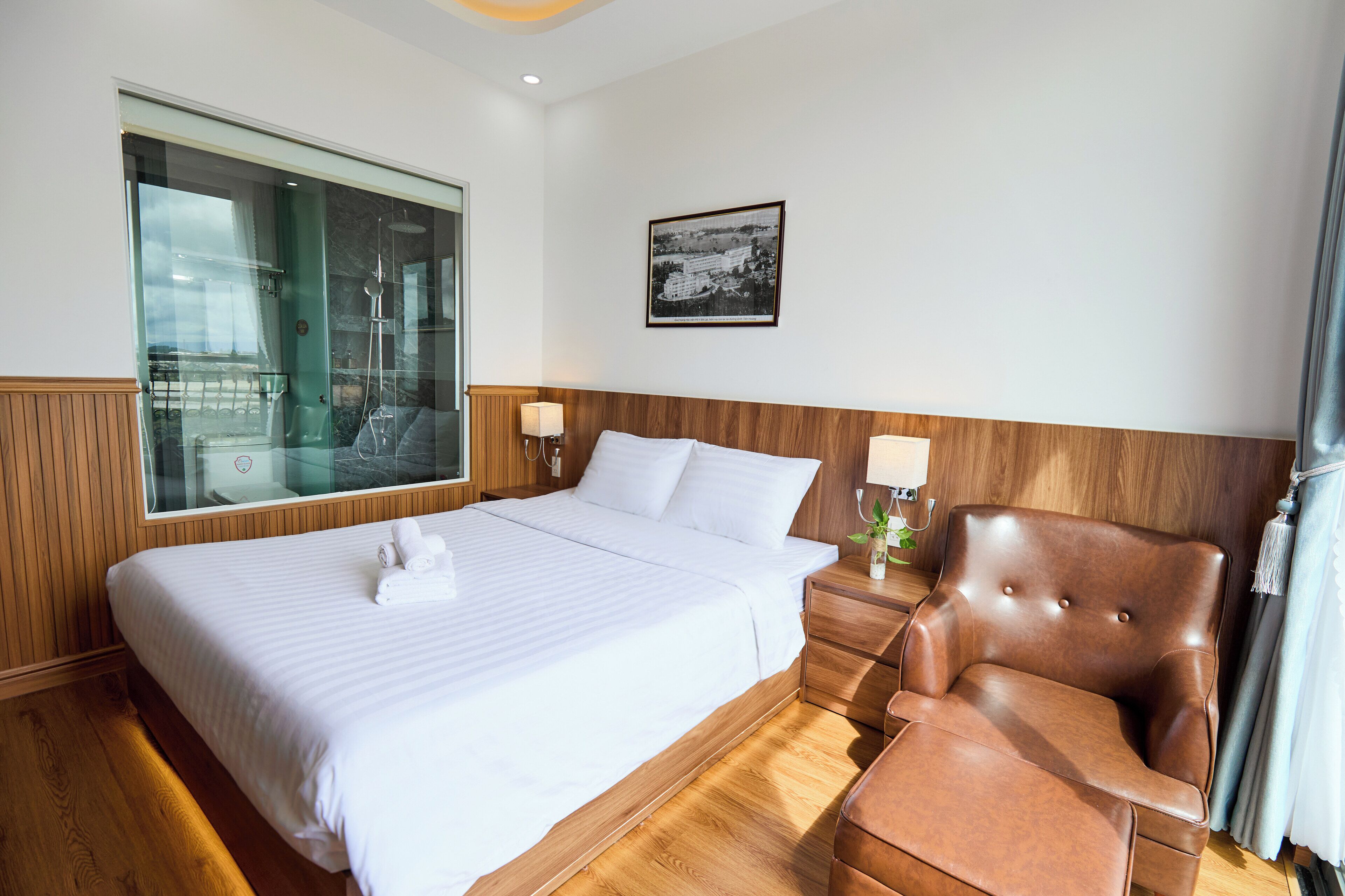 A hotel room with a large bed, wooden headboard, two bedside lamps, a brown leather armchair, and a large window with a view.