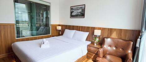 A hotel room with a large bed, wooden headboard, two bedside lamps, a brown leather armchair, and a large window with a view.