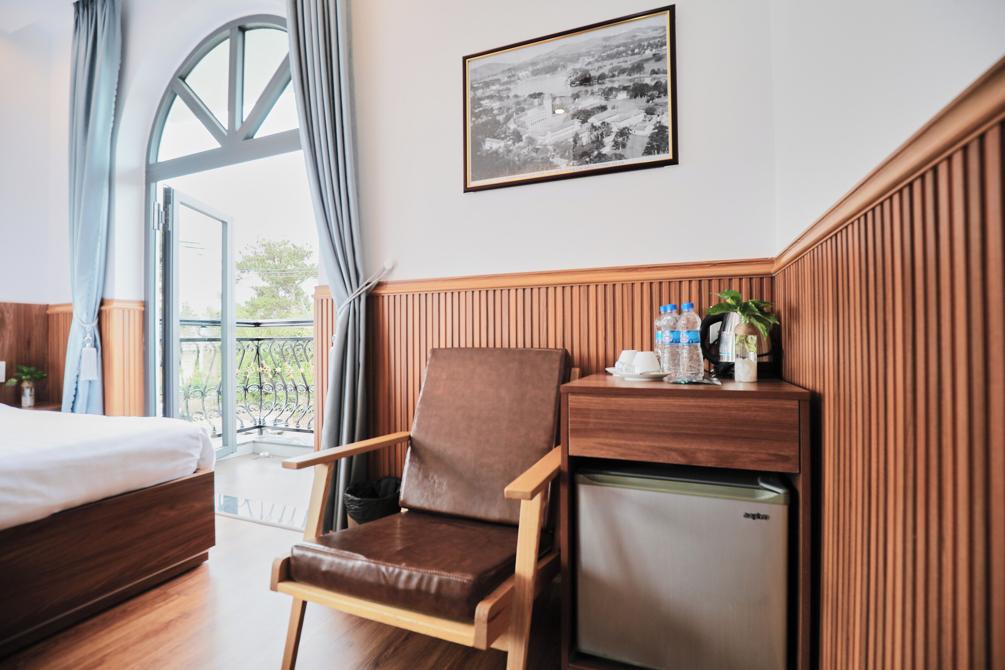Une chambre d’hôtel avec un lit, une chaise, un petit réfrigérateur, une fenêtre avec des rideaux et un tableau encadré au mur.