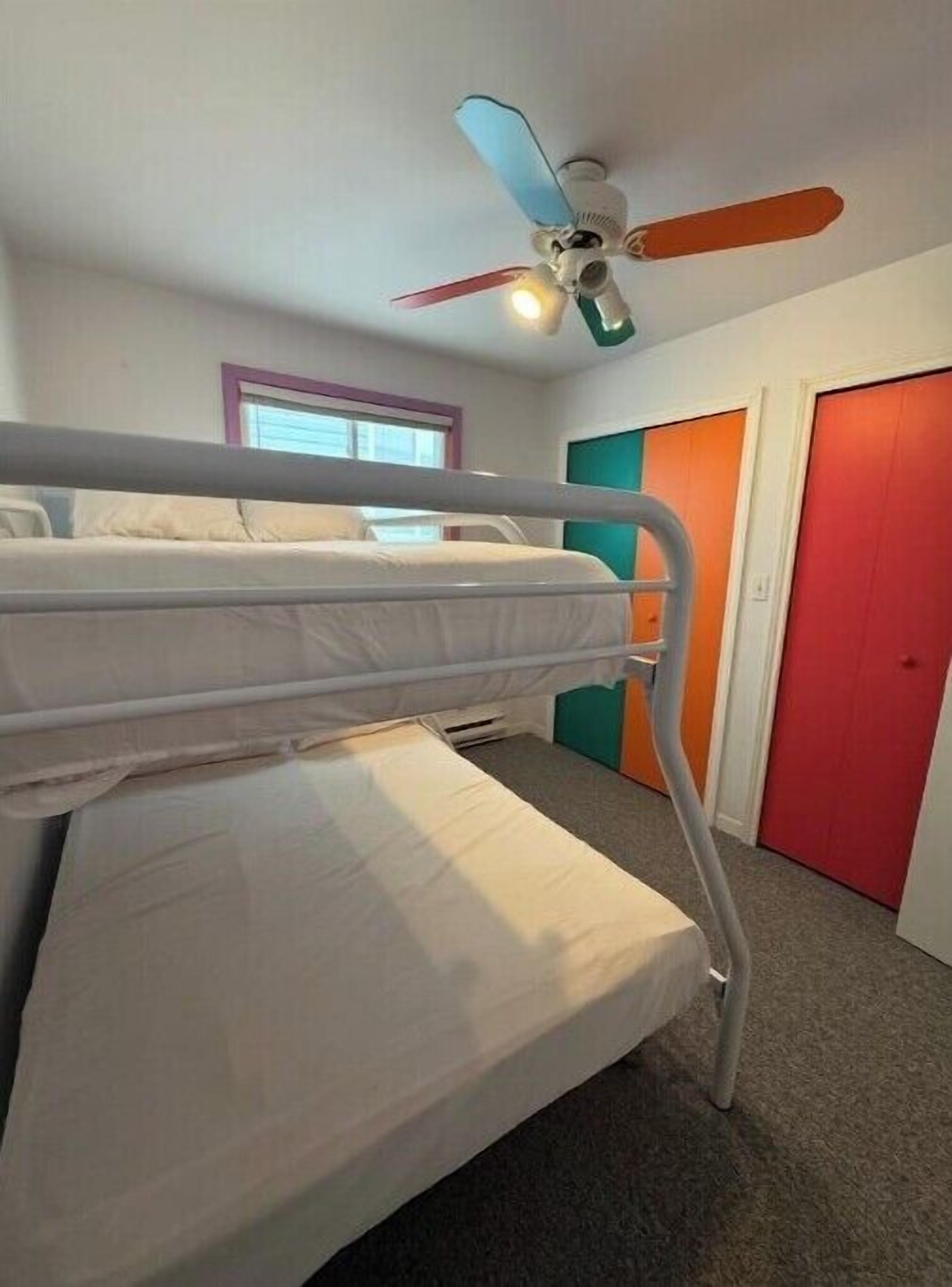 Een slaapkamer met stapelbed, plafondventilator, raam en deur met een kleurrijk paneel.