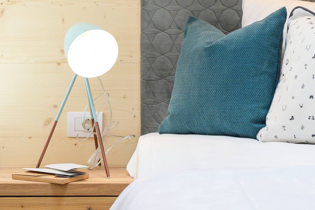 A neatly made bed with a white and patterned pillow, a teal pillow, and a round bedside lamp on a wooden nightstand.