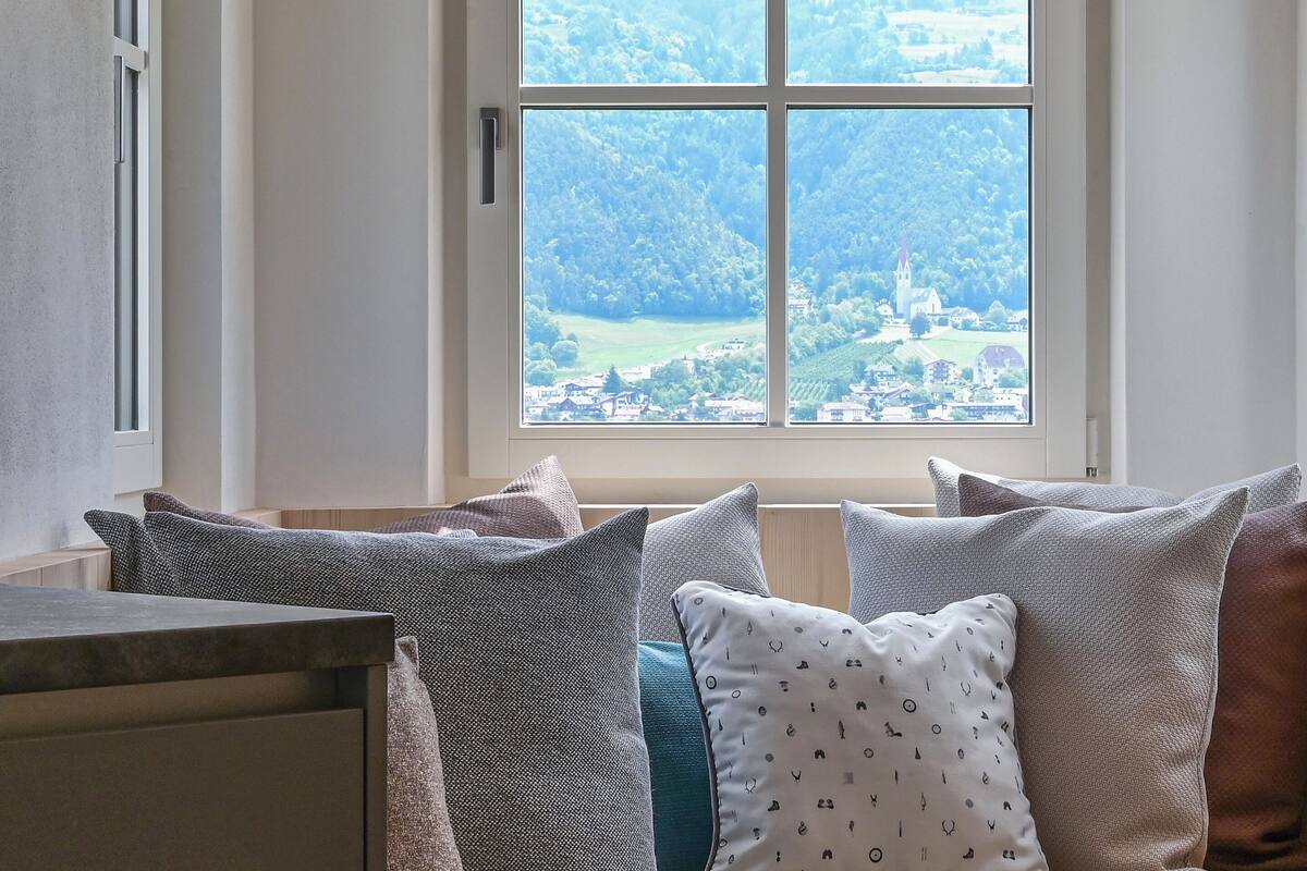 A room with a large window overlooking a village and hills. There are several pillows on a bench.