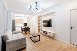 A modern living room with a grey sofa, wooden flooring, a TV mounted on a wall, and a dining area with chairs and a table.