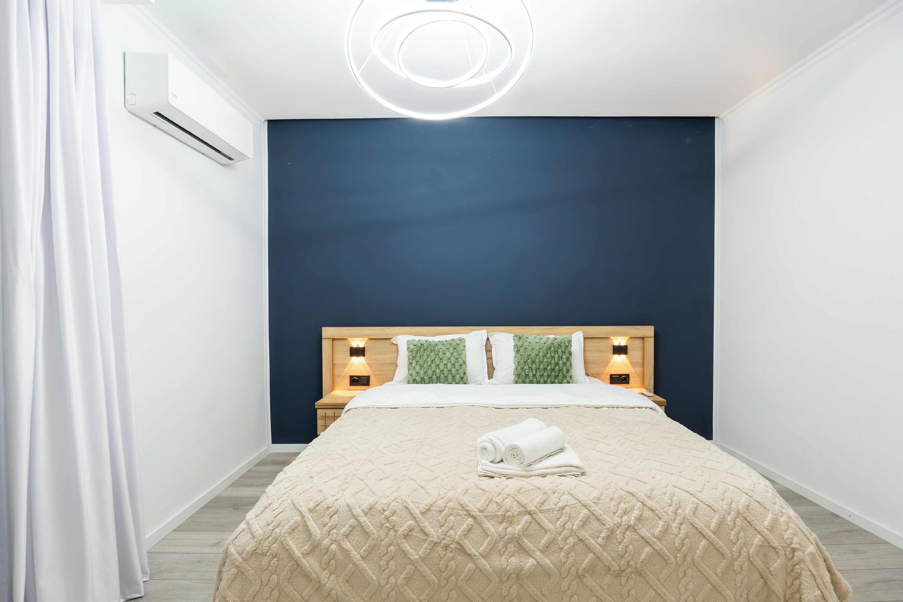 A modern hotel room with a large bed, a wooden headboard, two bedside tables with lamps, and a wall-mounted air conditioner.