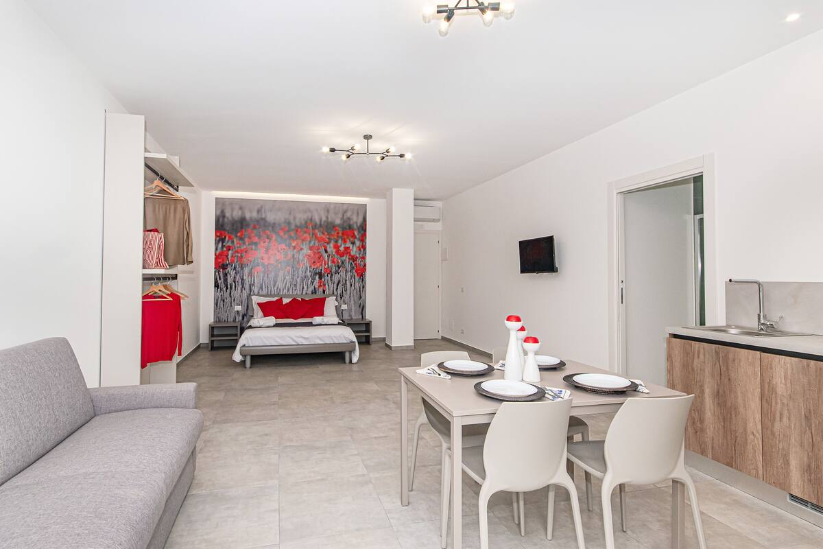 Une chambre d’hôtel moderne avec un coin repas, un lit agrémenté d’oreillers rouges, une armoire et une grande fenêtre ornée d’un tableau floral aux motifs rouges.