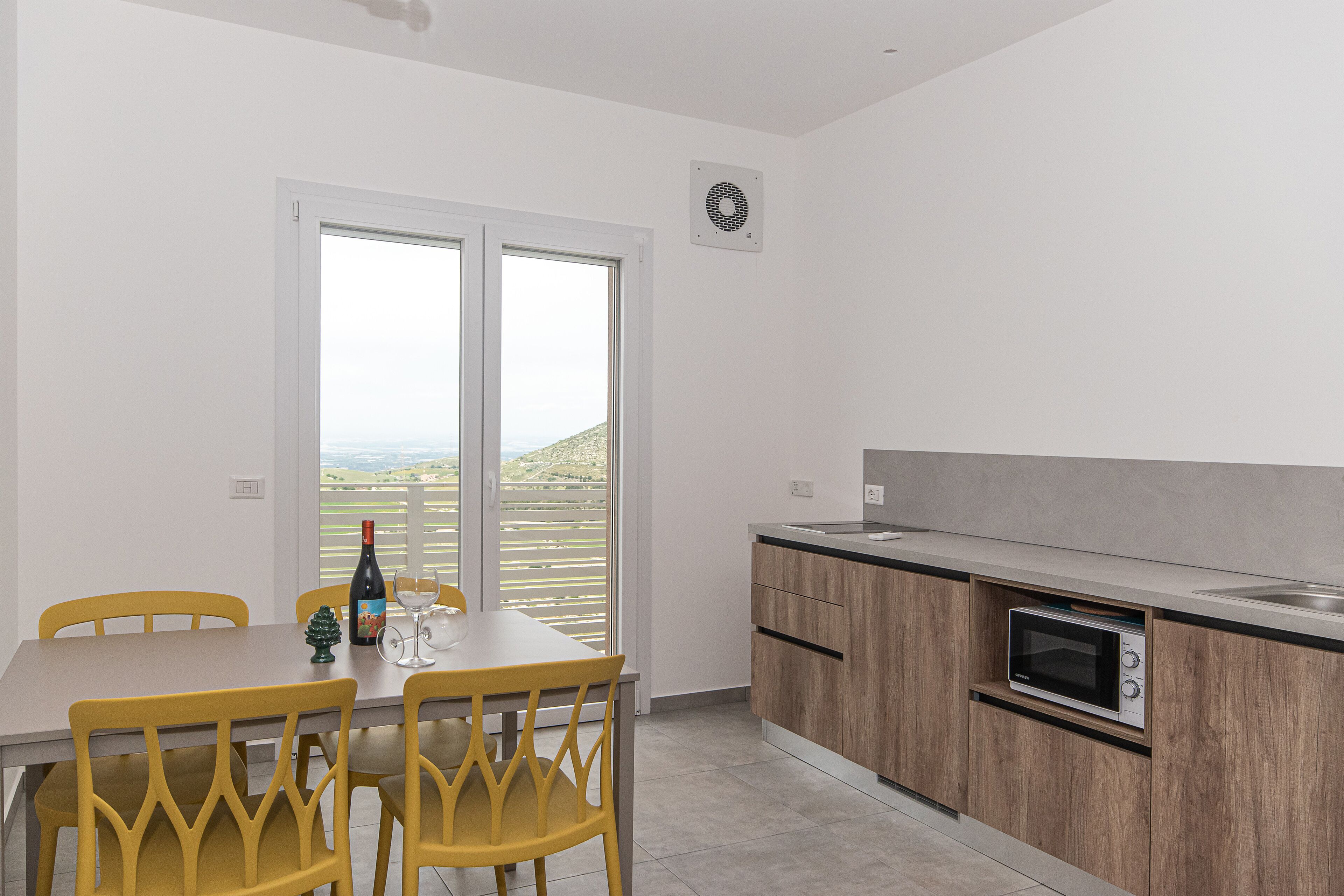 A modern kitchen with a built-in microwave and oven, a dining area with yellow chairs, and a large window offering a view of the countryside.