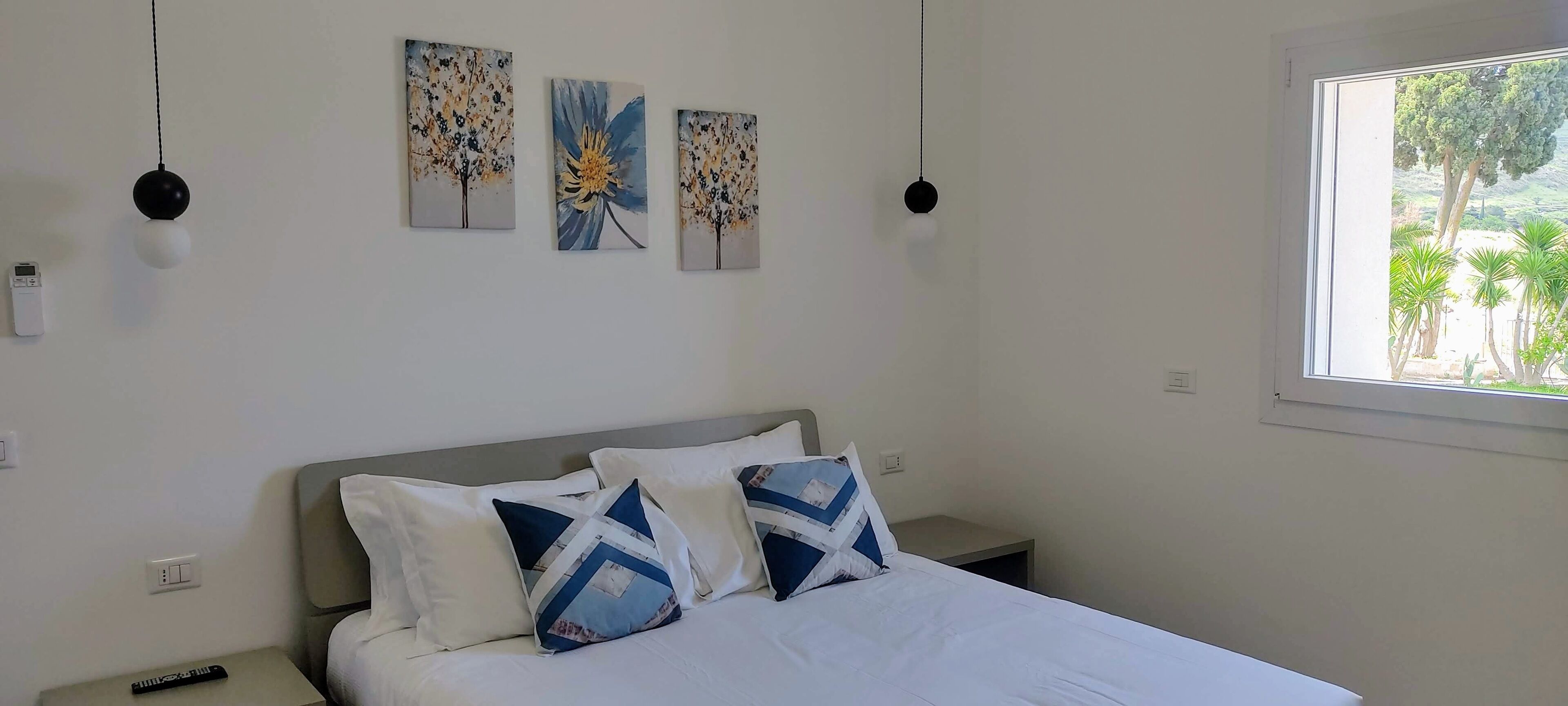 A bedroom with a bed, pillows, a nightstand, a remote control, and wall art.