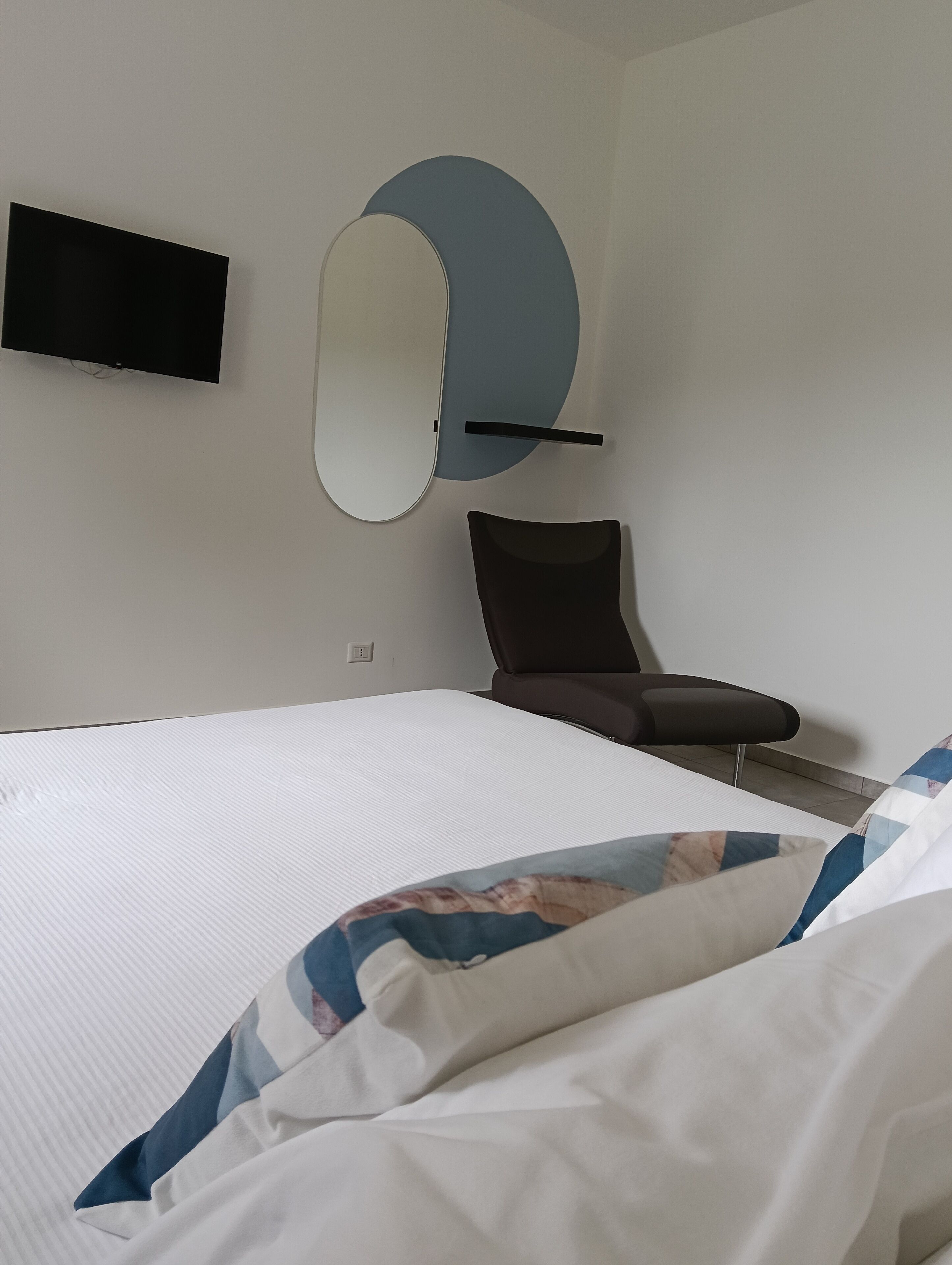 A hotel room with a bed, a chair, a TV mounted on the wall, and a round mirror.
