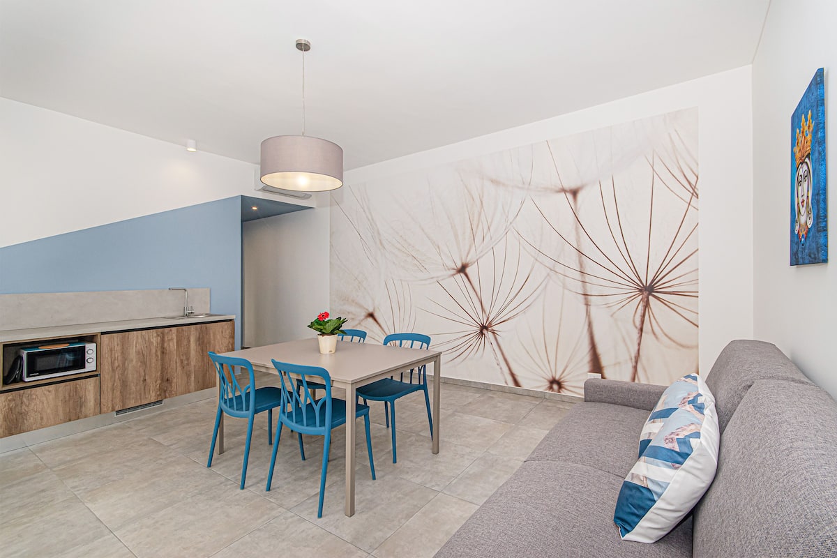 Un salon moderne comprenant un coin repas, un canapé gris, des chaises bleues et une table agrémentée d’un vase de fleurs.