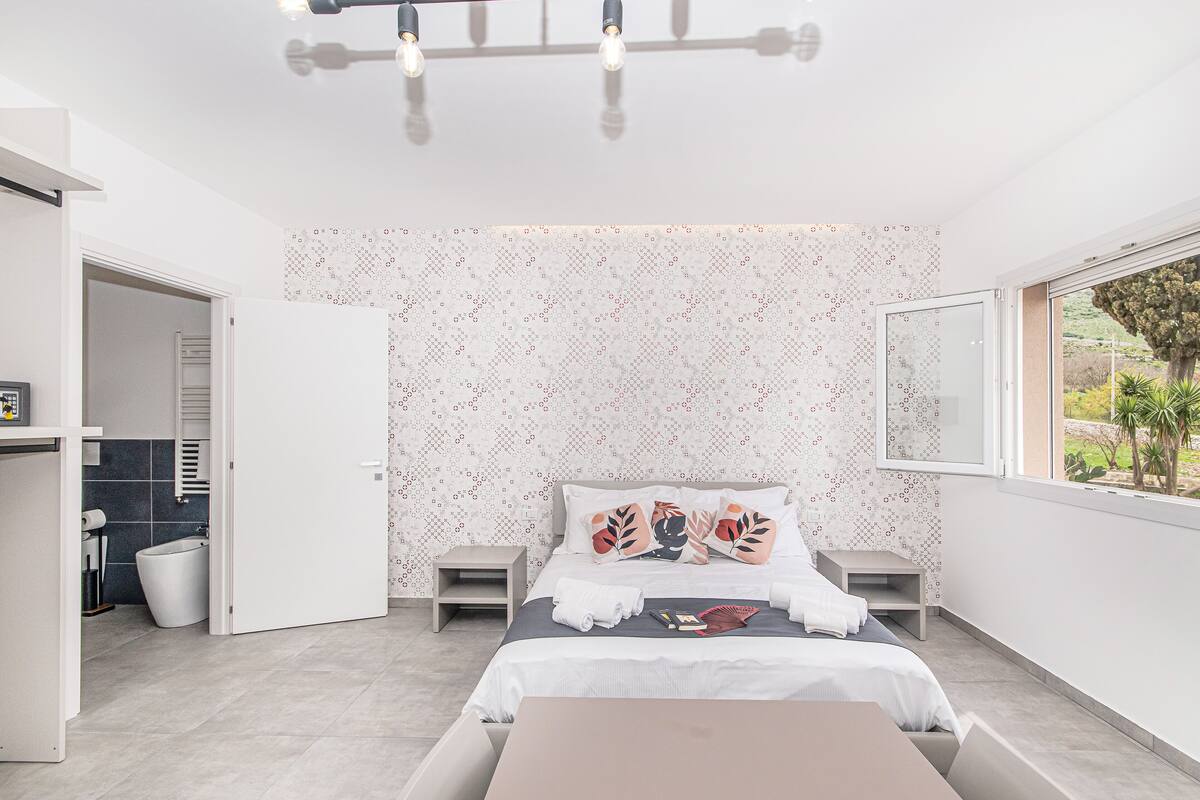 Une chambre à coucher moderne, avec un lit, des tables de chevet, une petite table d’appoint, un petit tabouret et une fenêtre donnant sur un paysage verdoyant.