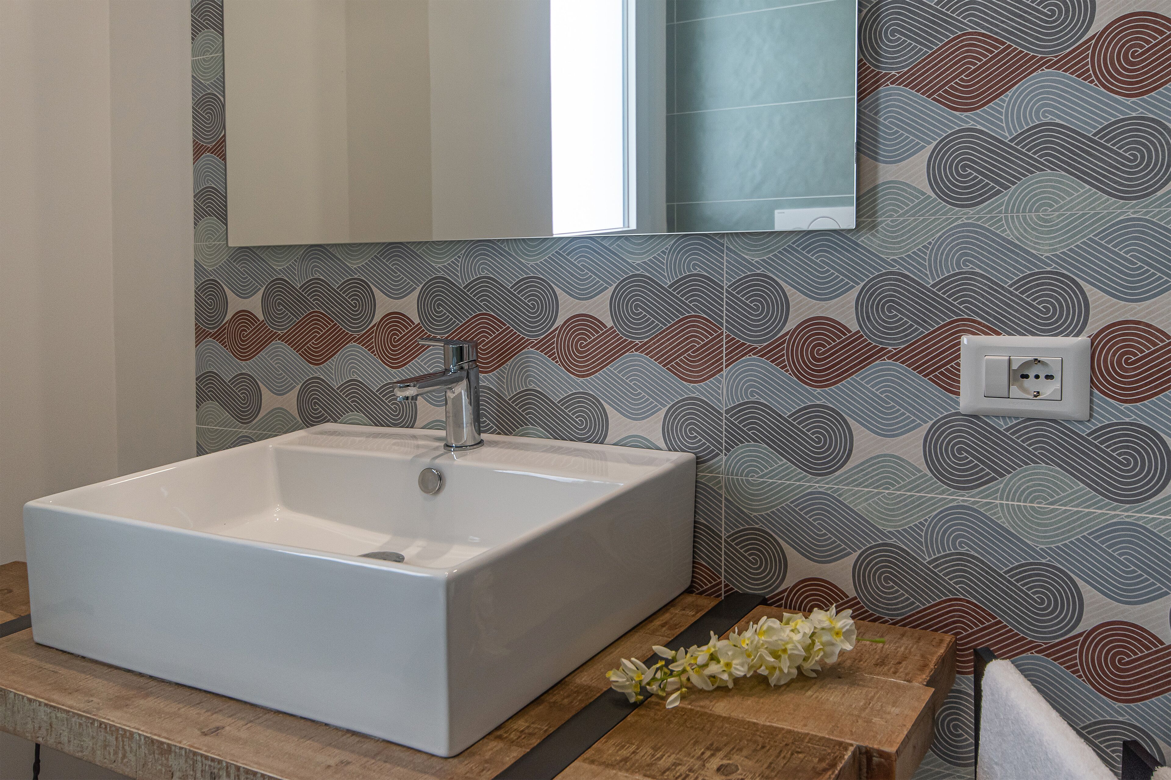 A bathroom with a rectangular white sink, a mirror above it, and patterned wallpaper.